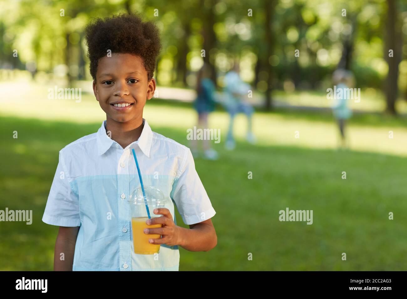 Waist-up-Porträt von lächelnden afroamerikanischen Jungen hält Orangensaft, während im grünen Park im Freien stehen, kopieren Raum Stockfoto