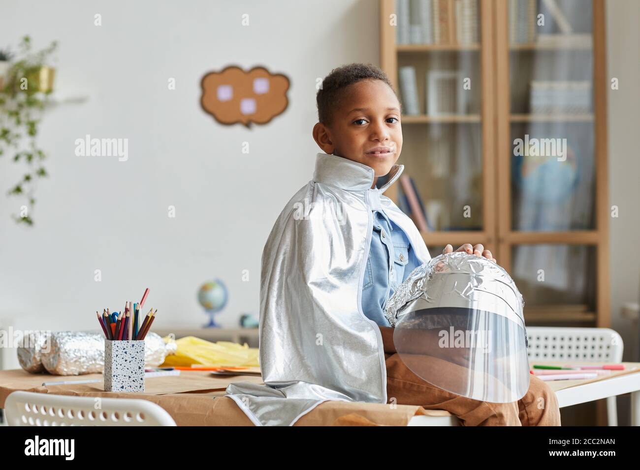 Portrait von niedlichen afroamerikanischen Jungen spielen Astronaut und Blick auf die Kamera, während genießen Kunst und Handwerk Unterricht in der Vorschule oder Entwicklungszentrum, Stockfoto