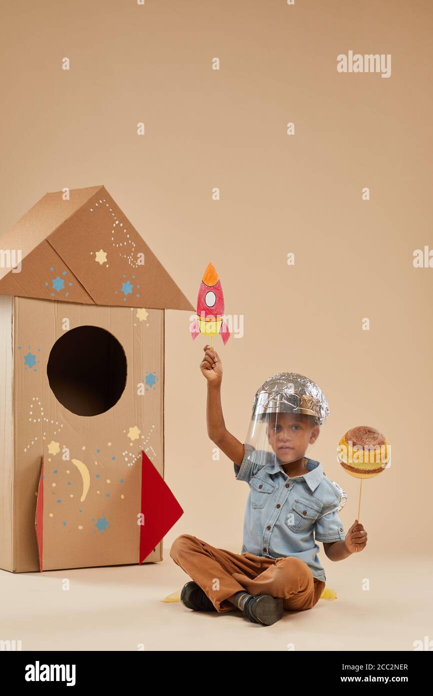 Vertikale voller Länge Porträt von niedlichen afroamerikanischen Jungen spielen Astronaut In handgefertigtem Raumanzug, während man auf dem Boden gegen Pappe sitzt Rakete Stockfoto