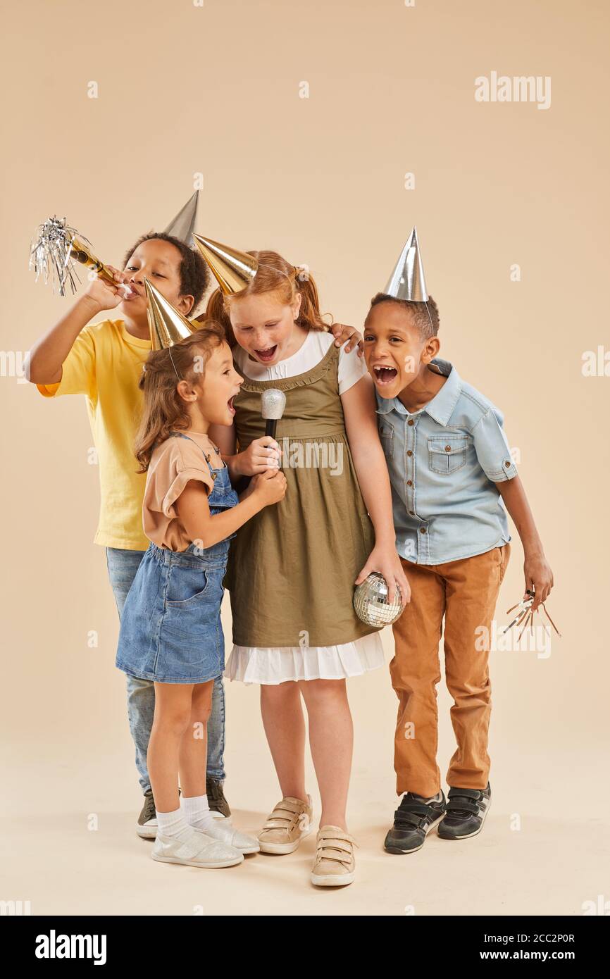 Vertikale voller Länge Porträt von verschiedenen Gruppe von Kindern tragen Partyhüte schreien zum Mikrofon, während sie vor schlichtem Hintergrund stehen In studi Stockfoto