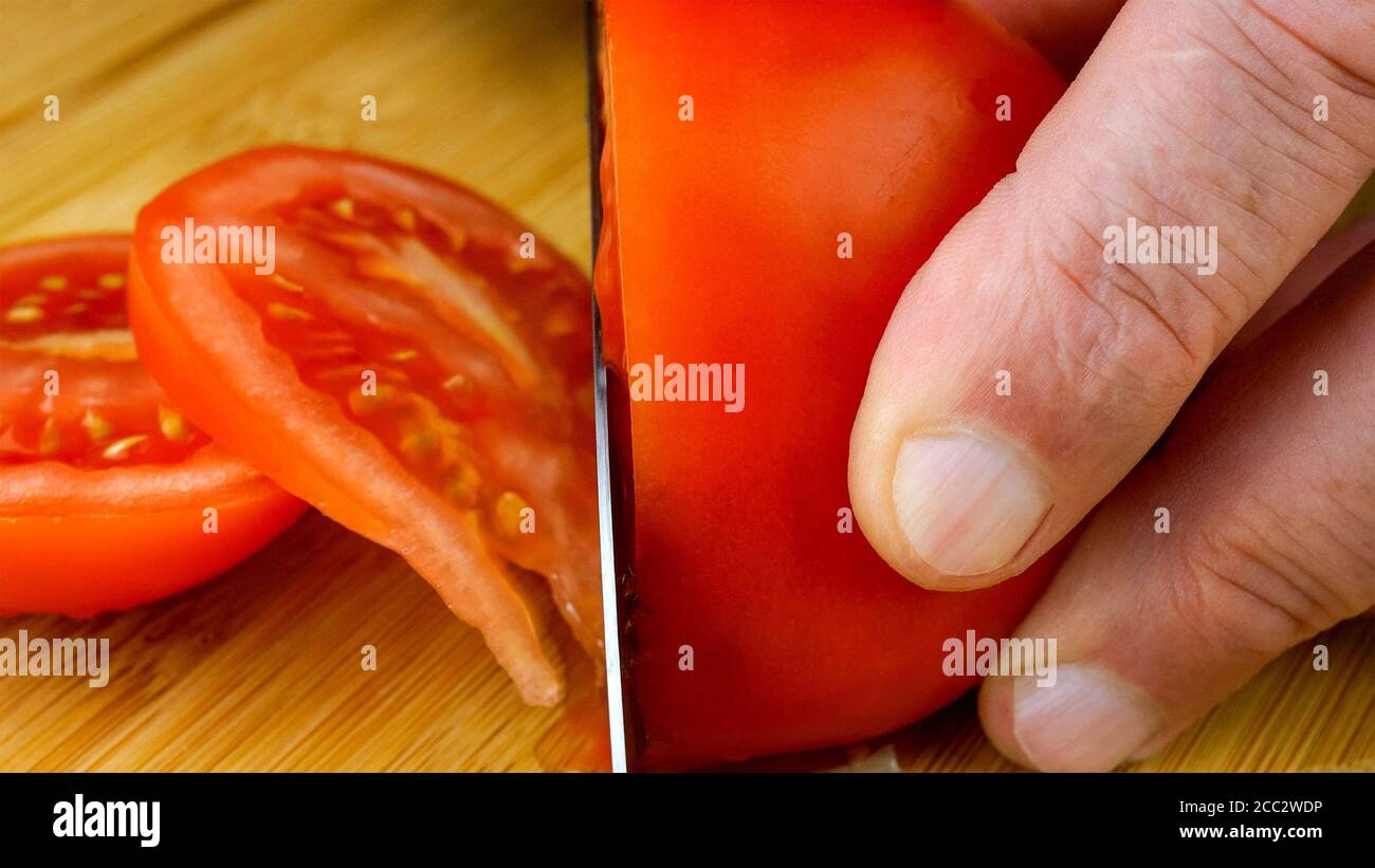 Die Hände des Küchenchefs schneiden saftige reife Tomaten mit Küchenmesser auf Holzschneidebrett. Nahaufnahme. 16x9-Format. In Innenräumen. Stockfoto