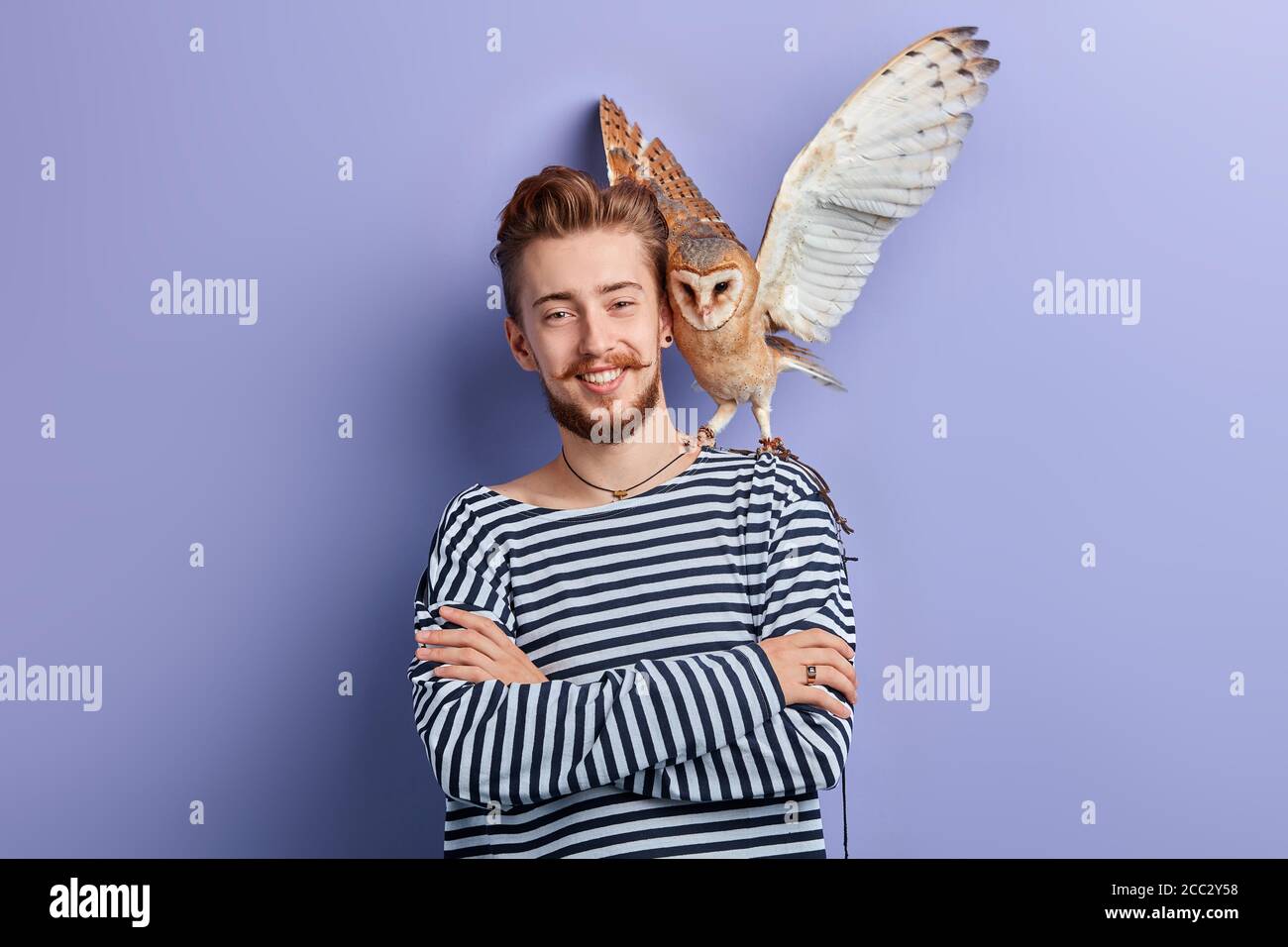 Fröhlicher positiver junger Mann mit einer Eule in der Schulter, die fliegen wird. Isolierter blauer Hintergrund, Studioaufnahme. Stockfoto