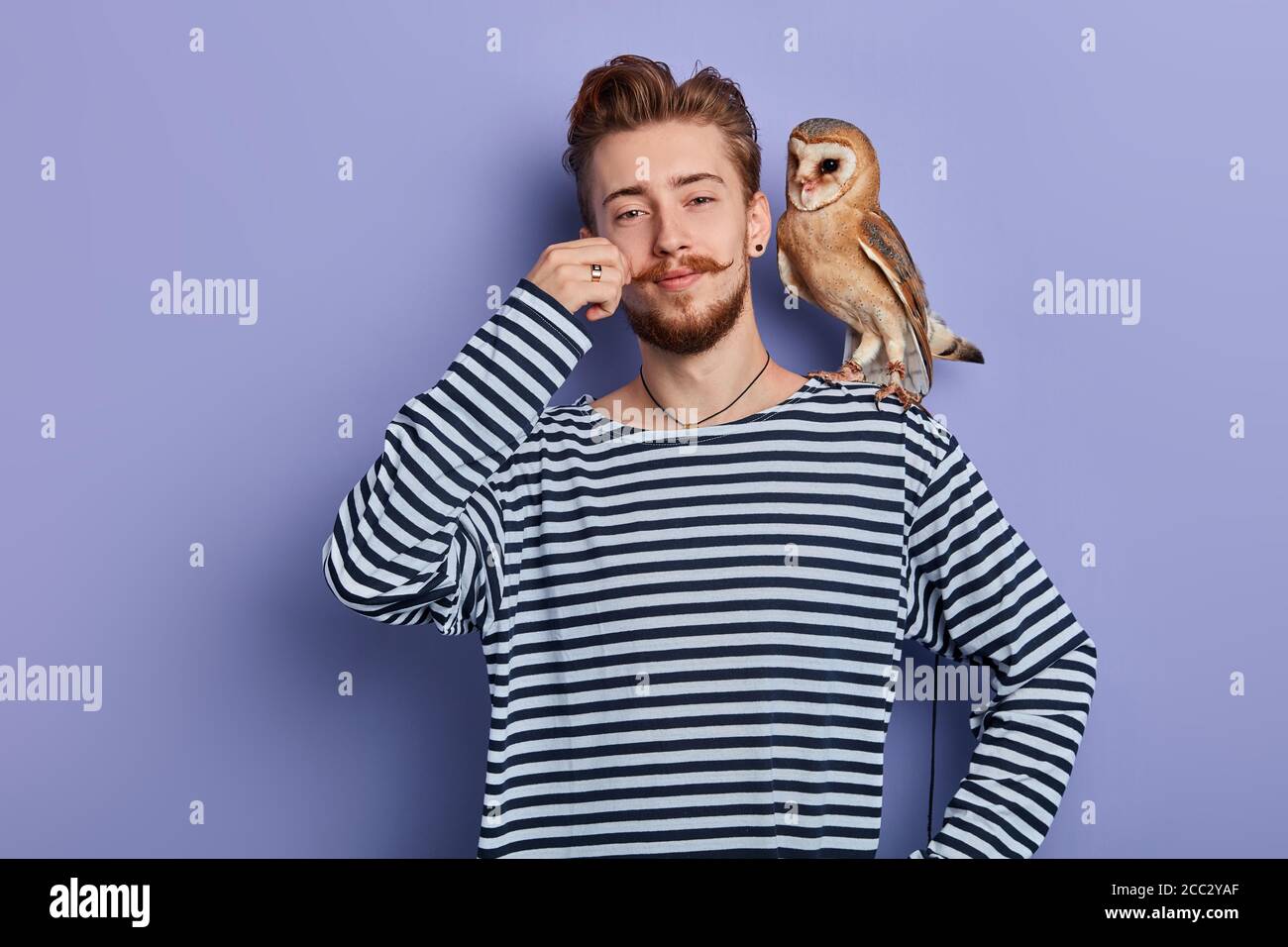 Fröhlicher lustiger schlauer Kerl im gestreiften Pullover, der Geld vom Touristen nimmt, weil er ein Pictyre mit einer Eule nimmt, isolierter blauer Hintergrund, Studioaufnahme, Busin Stockfoto