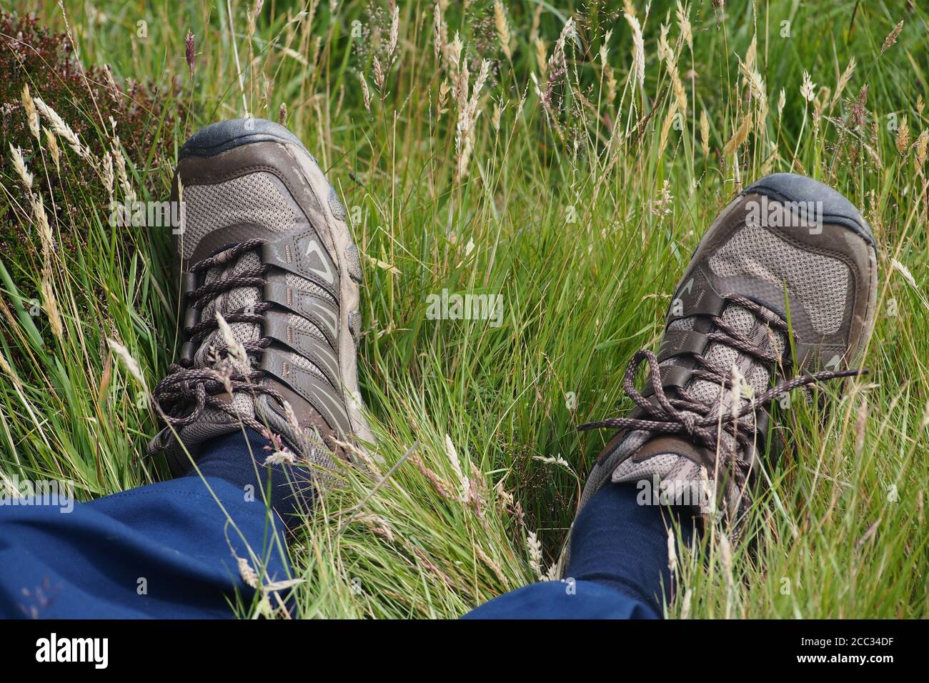 Ein Blick auf die Knöchel und Füße eines Mannes, der in rauem Gras liegt und blaue Hosen, blaue Socken und Wanderschuhe trägt Stockfoto