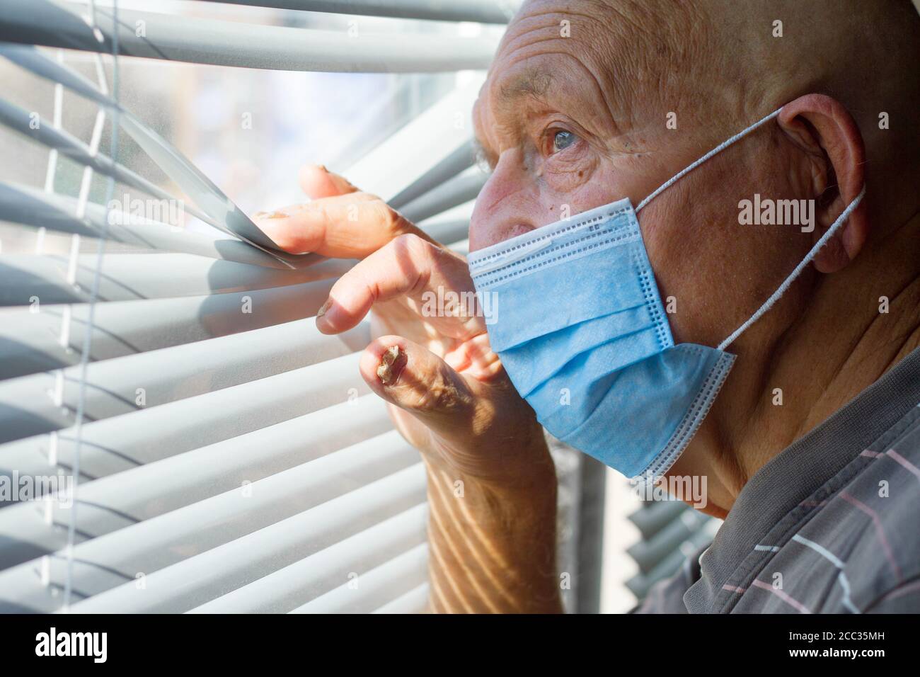 Sehr älterer Mann in blauer Schutzmaske auf seinem Gesicht schaut durch das Fenster, zu Hause bleiben Konzept Stockfoto