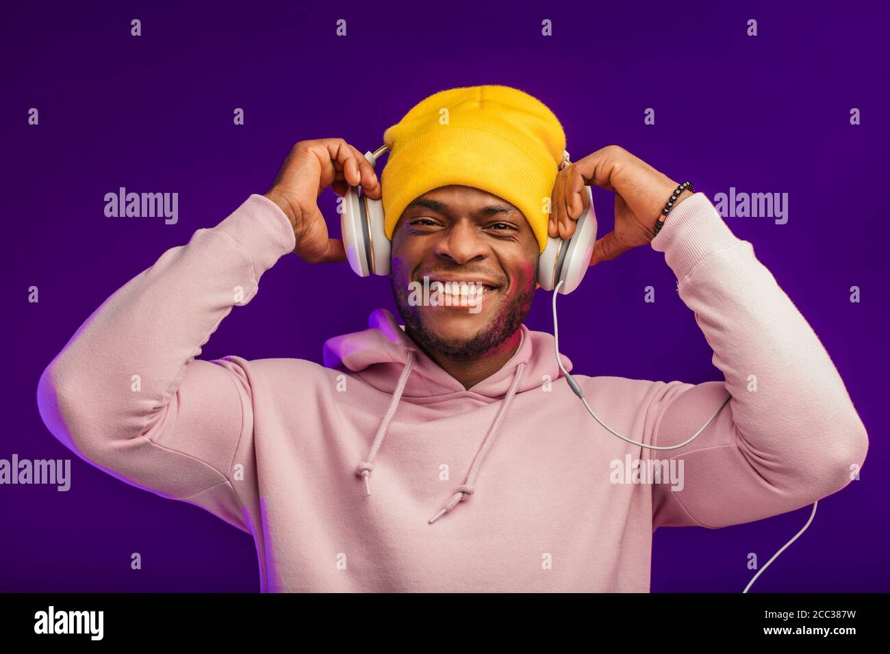 Junge afrikanische Mann in Hoodie, in der Sinn für Mode eines street Urchin, drehte sich ein wenig in der Kopfhörer auf der violette Hintergrund isoliert, holding Ohrhörer Stockfoto