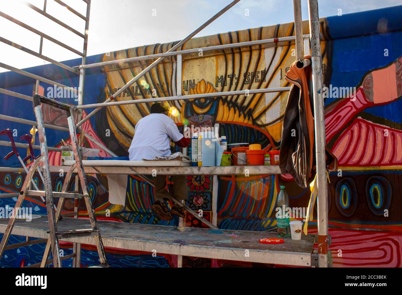 Berlin, Deutschland 09/14/2009: Ein älterer Mann sitzt auf einem Arbeitsgerüst voll von Malausrüstungen und malt ein Bild auf Berliner Mauer mit hallo Stockfoto
