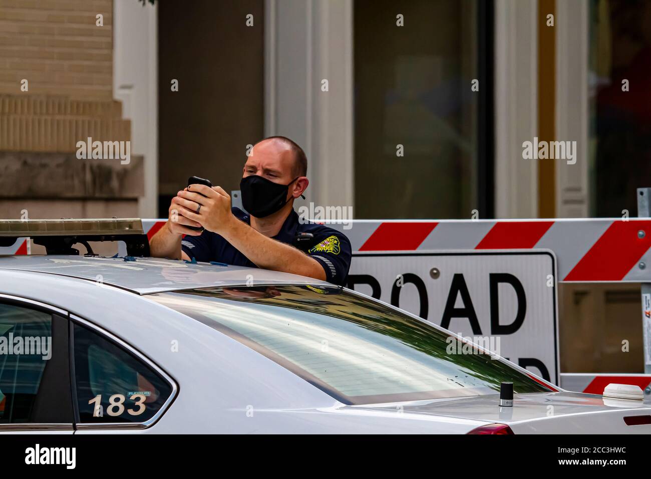 Frederick, MD, USA 08/14/2020: Ein junger kaukasischer Polizist trägt im Dienst eine Gesichtsmaske. Er steht bei seinem Polizeiwagen vor der Tür Stockfoto