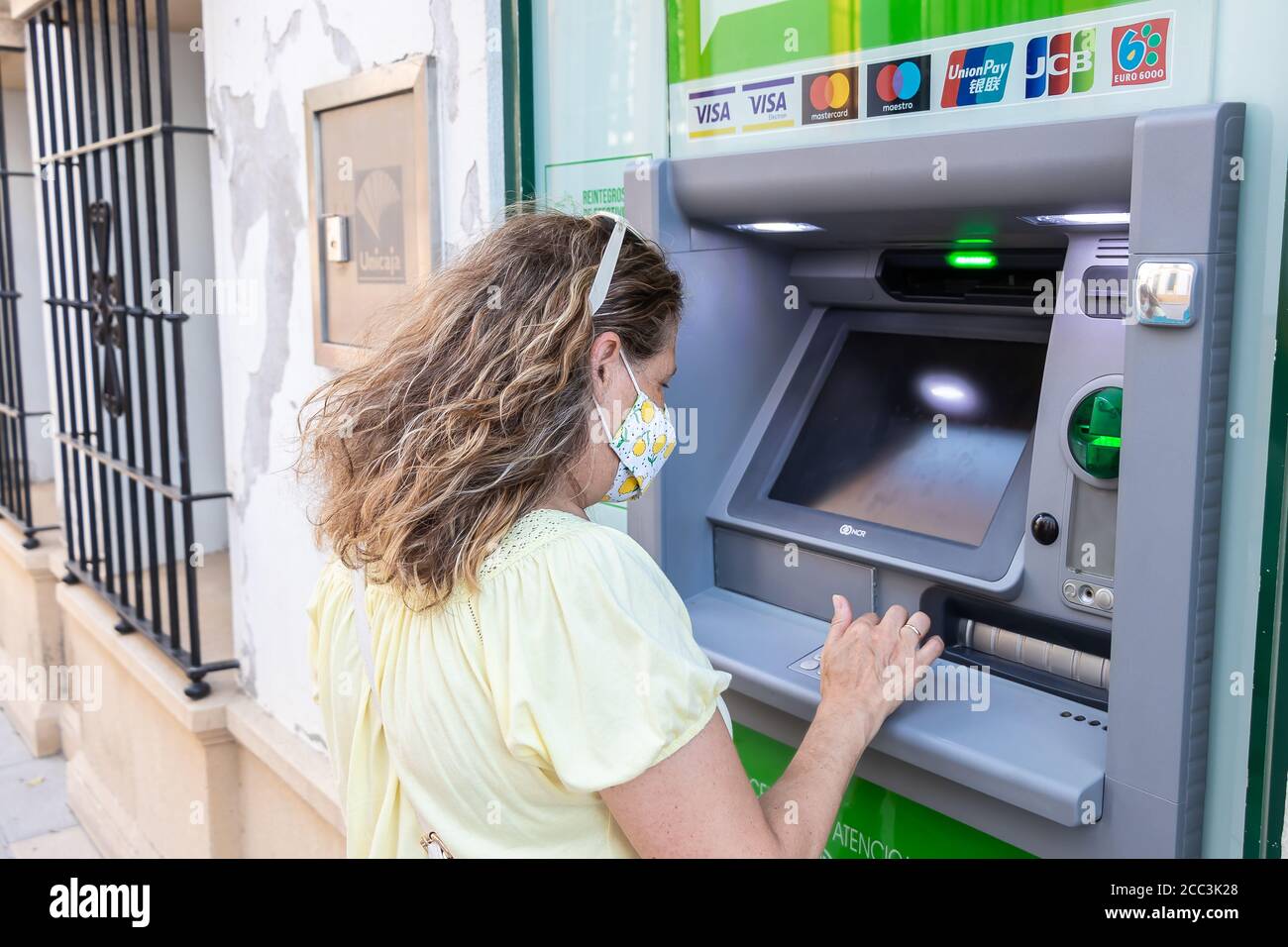 Huelva, Spanien - 16. August 2020: Frau in Schutzmaske benutzt einen Geldautomaten. Neue Normalität in Spanien durch Covid-19 Coronavirus Stockfoto