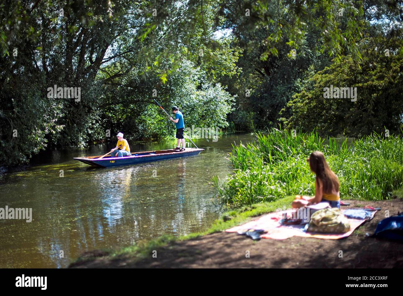 Ein Punt-Paar passiert eine Frau, die neben dem River Cam in Grantchester bei Cambridge sitzt, während das warme Wetter weitergeht. Stockfoto