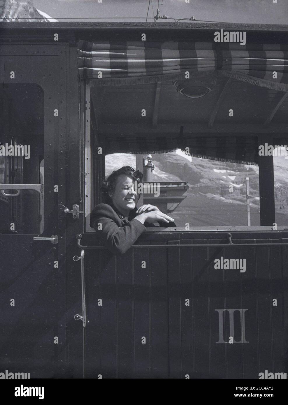 1950s, historisch, eine Frau, die in einem Schweizer Bergbahnwagen sitzt und sich aus einem offenen Fenster lehnt, Schweizer Alpen, Schweiz. Die Zahl 3 in römischen Ziffern steht in Buchstaben auf der Waggontür. Stockfoto