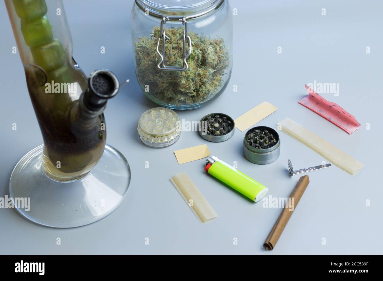 Rauchen von Cannabis Zubehör und Sachen auf grauem Hintergrund. Bong, stumpfes und gemeinsames Papier, Mahlwerk und Marihuanaknospen im Glas Stockfoto