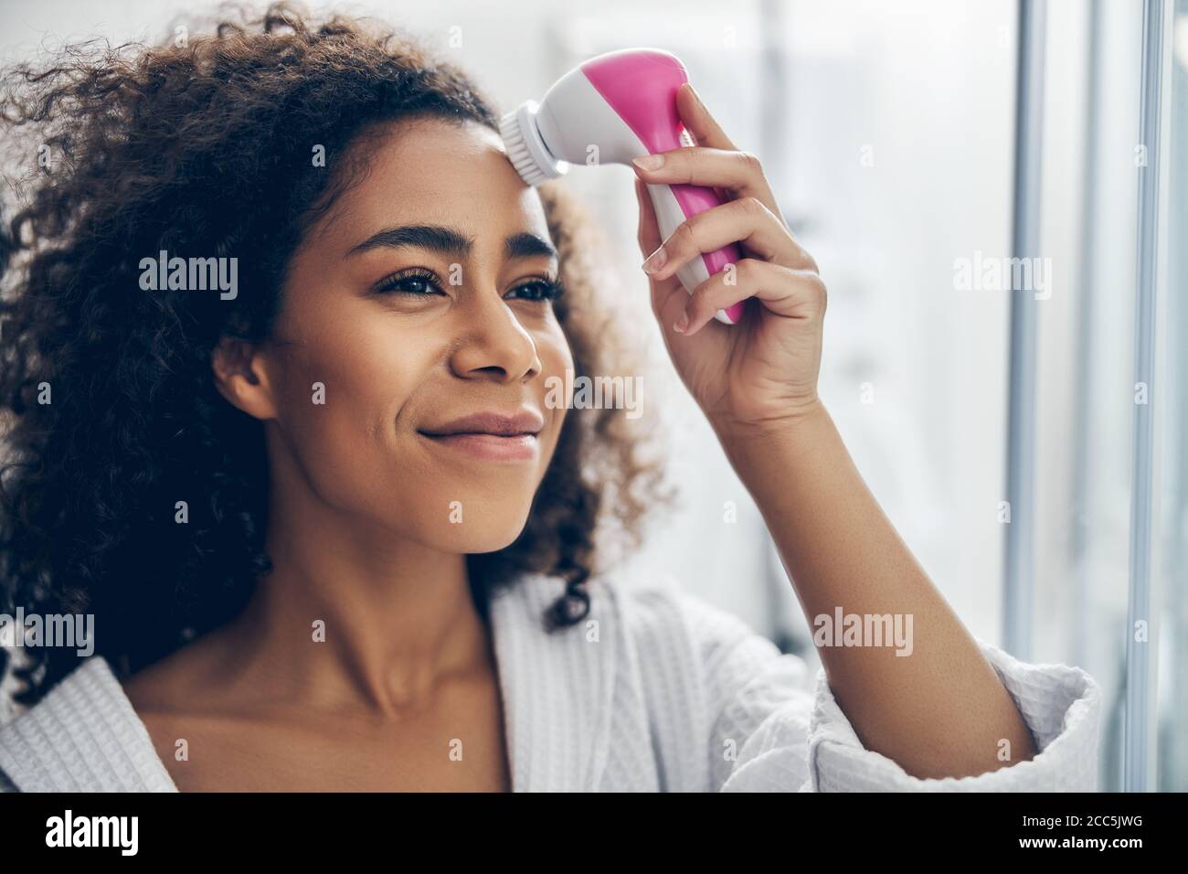 Verträumte Frau, die eine elektrische Gesichtsreinigungsbürste anwendet Stockfoto