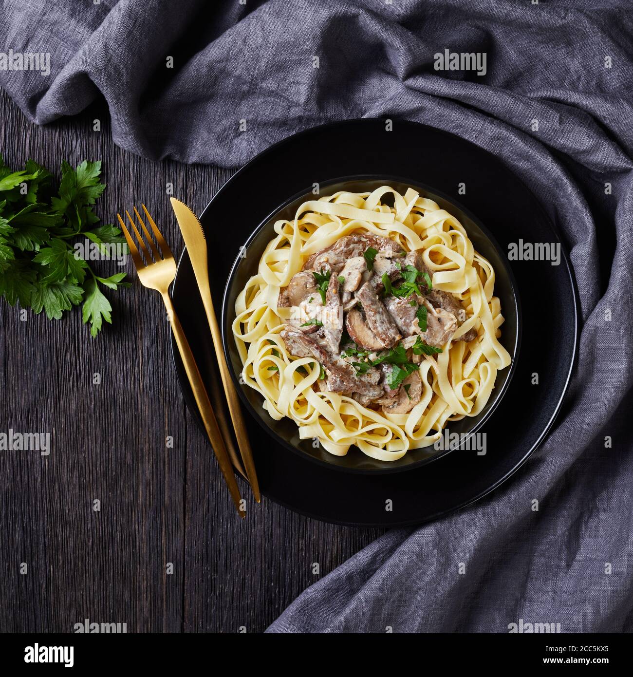 Rind Stroganoff gekocht mit saurer Sahne Pilze Sauce, serviert mit Tagliatelle in einer schwarzen Schüssel mit goldenen Gabel und Messer auf einem dunklen Holztisch, Klo Stockfoto