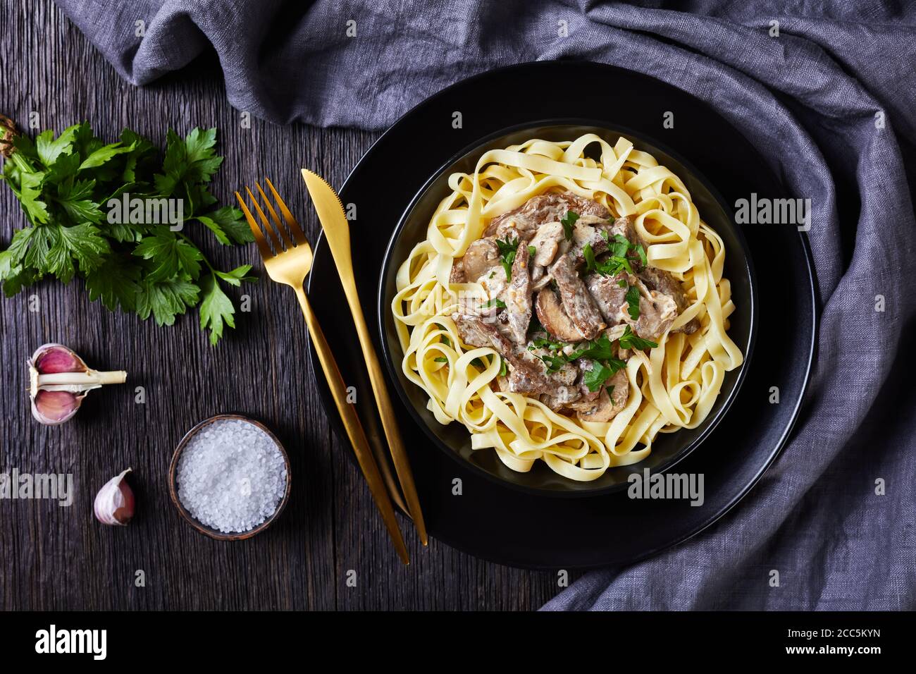 Rindfleisch Stroganoff gekocht mit saurer Sahne Pilze Sauce, serviert mit Tagliatelle in einer schwarzen Schüssel mit goldenen Gabel und Messer auf einem dunklen Holztisch, Fla Stockfoto