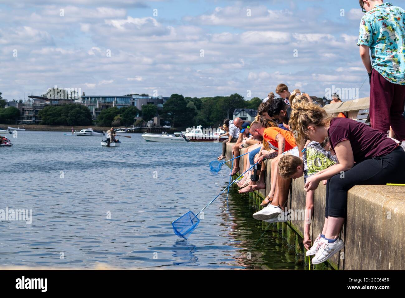 Mudeford Quay, Vereinigtes Königreich - 16 July 2020: Viele Menschen sitzen in einer Reihe auf Beton Ufer Linie fangen Krabben zu Krabbenfallen und Netze, Familien Tag Stockfoto