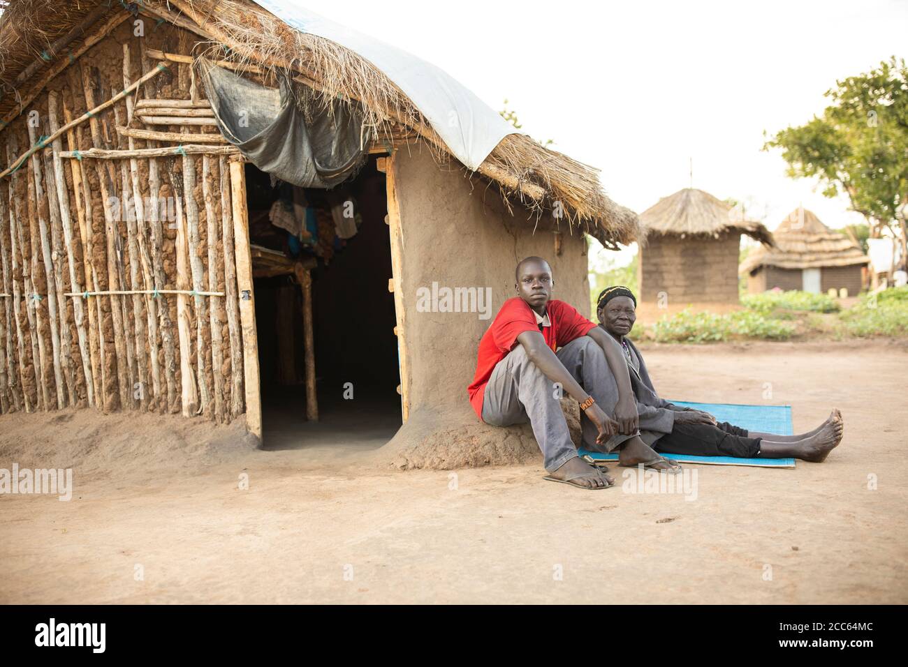 Ein junger Mann und seine alte Großmutter sitzen vor ihrem Haus zusammen in der Palabek Flüchtlingssiedlung im Norden Ugandas, Ostafrika. Stockfoto