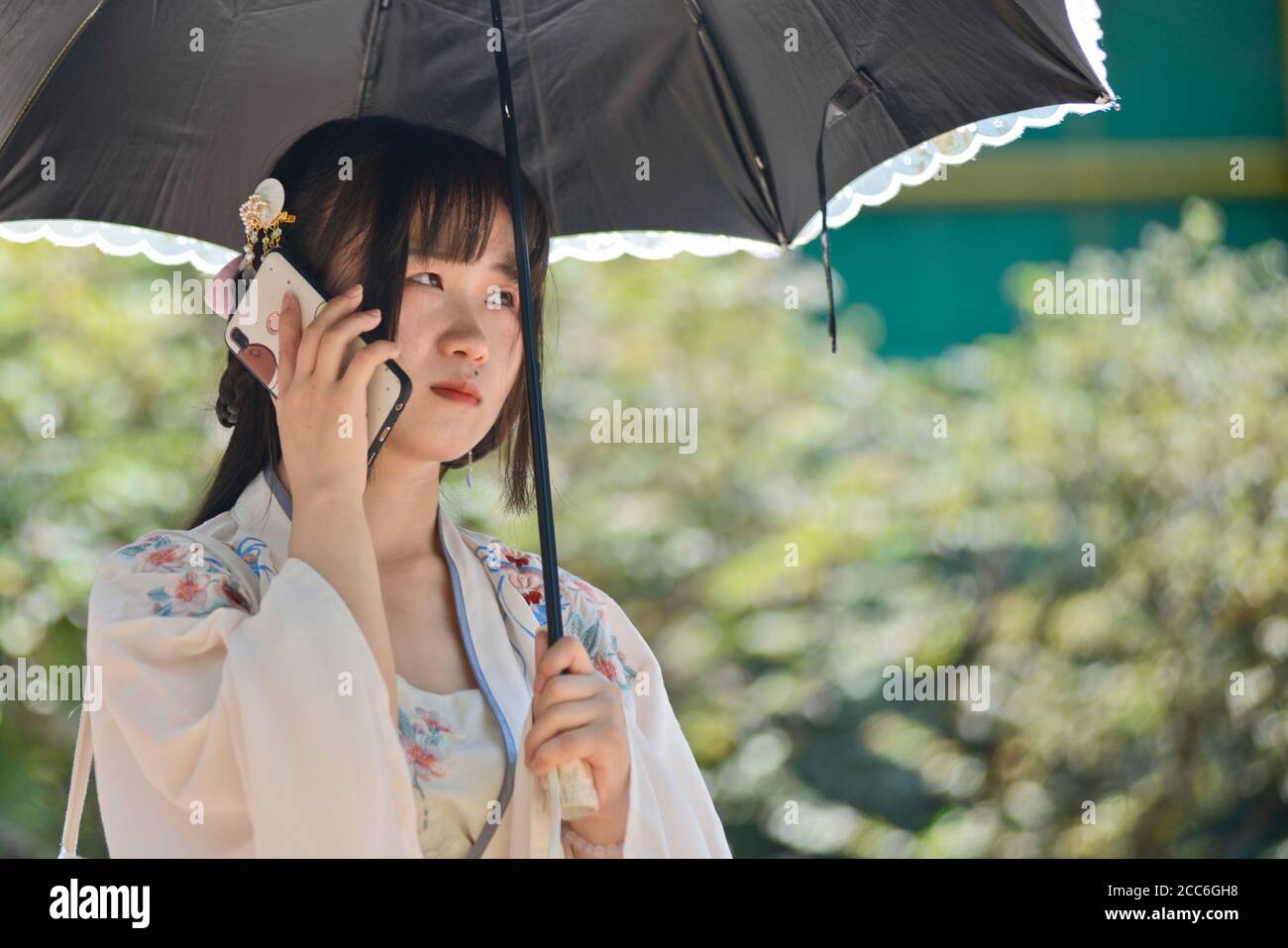 Ein chinesisches junges Mädchen, das mit traditioneller Kleidung und Regenschirm gekleidet ist und mit ihrem Handy spricht. Jianghan Road, Wuhan, China Stockfoto