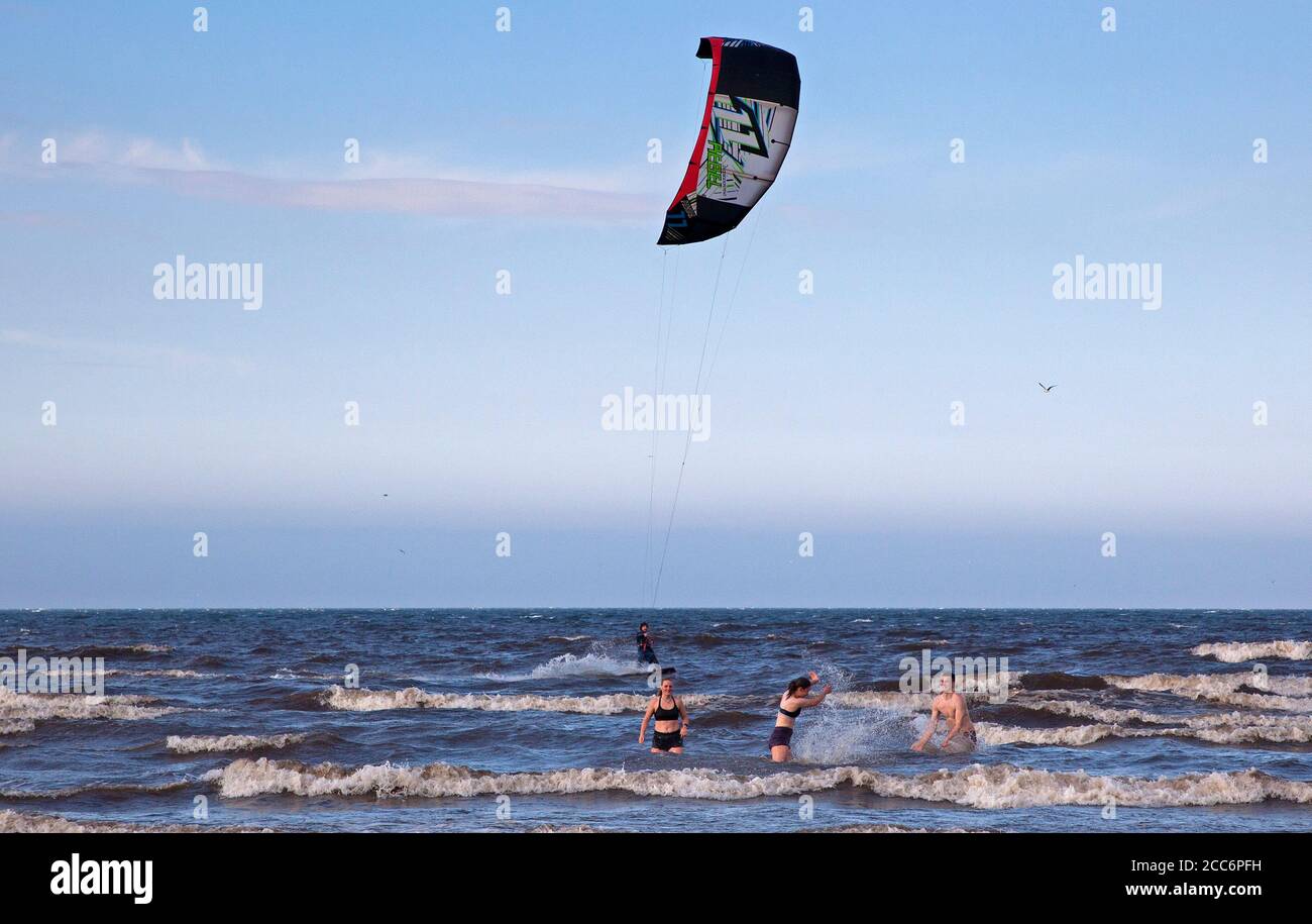 Portobello, Edinburgh, Schottland, Großbritannien. 19. August 2020. Windiger Nachmittag blies den Dunst weg, der am Morgen mit verstreuten Sonnenstrahlen später anhielt. Wind 22 km/h mit möglichen Böen von 42 km/h, die diesem Kite Surfer helfen, ein paar Airs zu bekommen. Im Bild: Männliche und zwei weibliche Badegäste, die im Vordergrund herumplanschen. Stockfoto