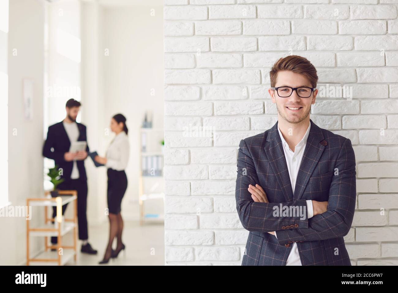 Lächelnder positiver Geschäftsmann in legerer Kleidung, der in einem weißen Büro an einer weißen Wand steht. Stockfoto