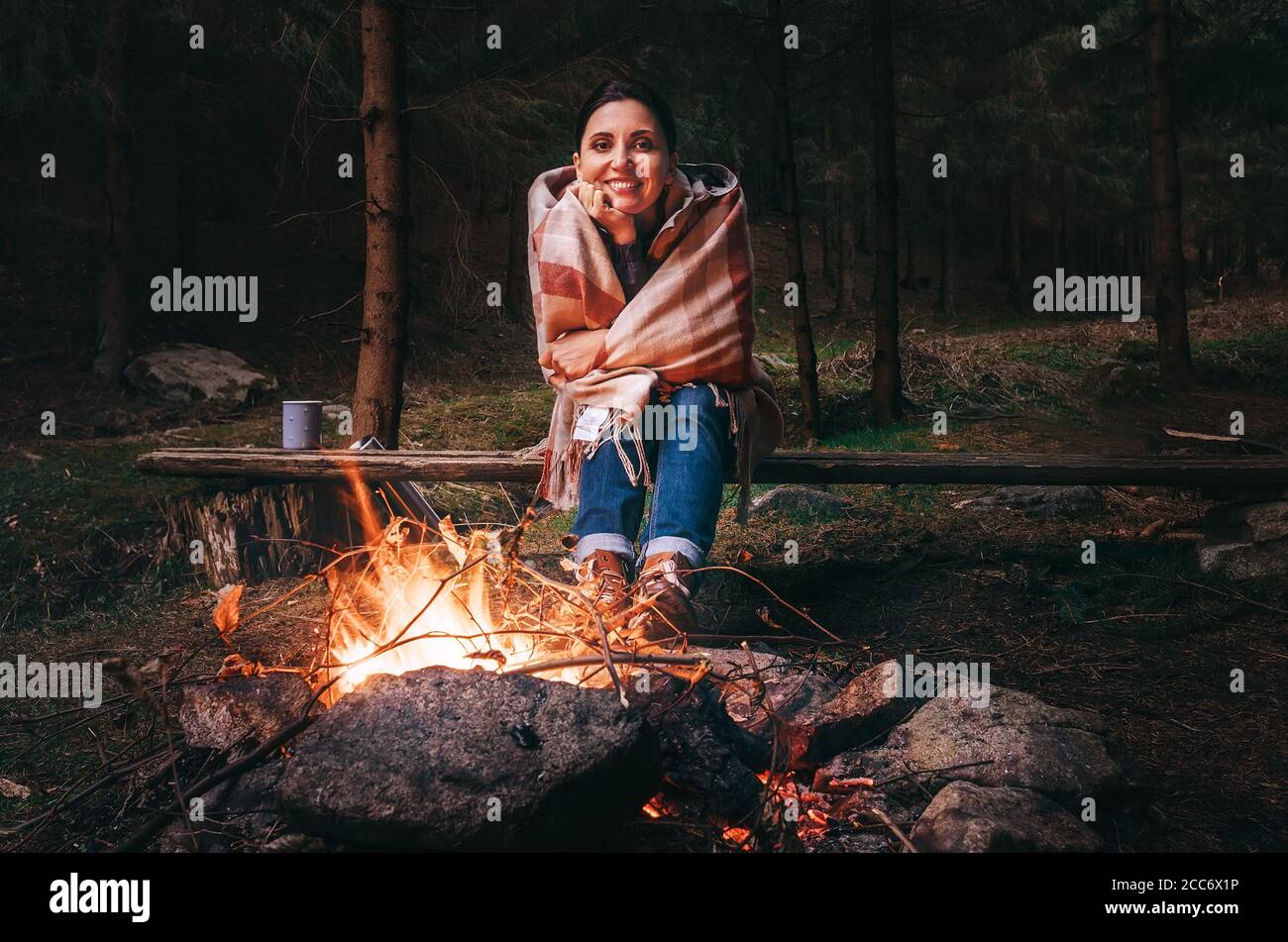 Hübsche lächelnde junge Frau sitzt in der Dämmerung am Lagerfeuer Herbstwald Stockfoto