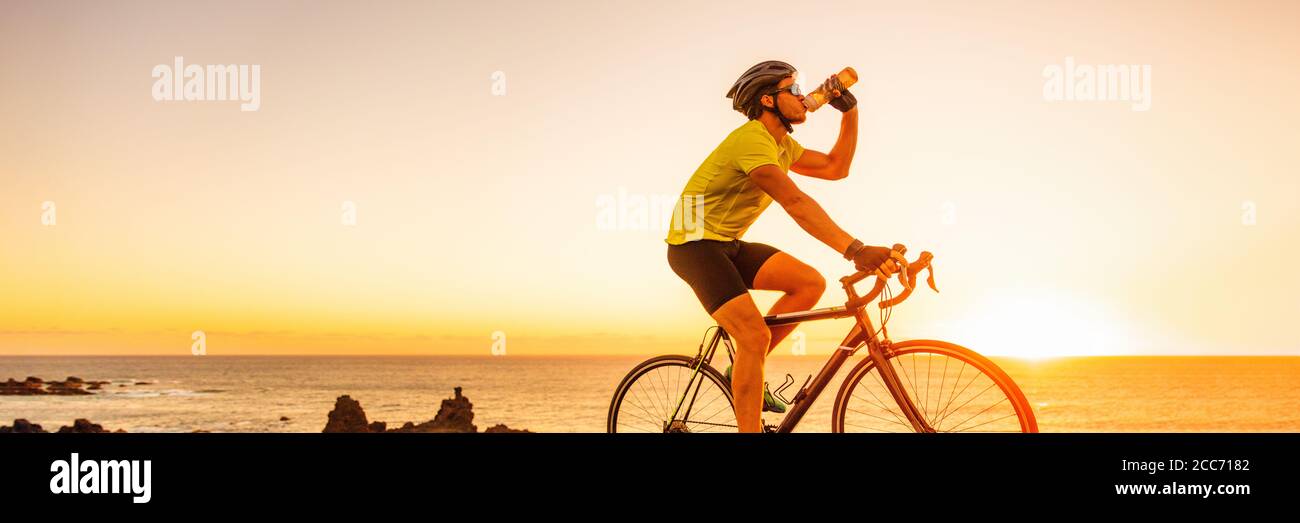 Triahtlon Athlet Mann Trinkwasser Flasche auf Straße Rennrad Fahrt im Freien bei Sonnenuntergang Banner Panorama-Landschaft. Radfahrer Radfahren draußen mit Stockfoto