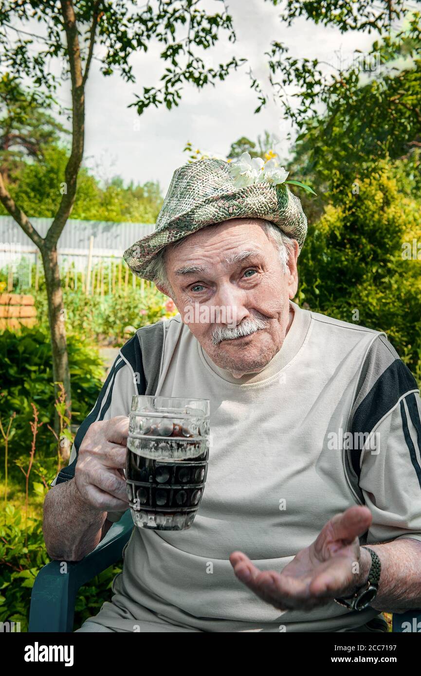 Porträt eines hübschen älteren Mannes von 87 Jahren, der nach der Arbeit im Garten ein kühles Getränk trinkt. Glückliches aktives Alter. Ein Mann trinkt dunkles Bier Stockfoto