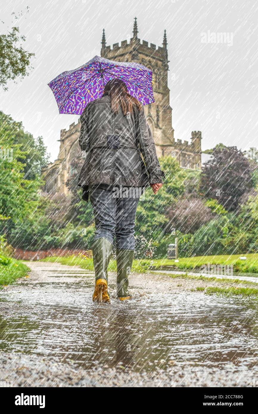 Rückansicht einer Frau, die Gummistiefel trägt und einen Regenschirm trägt, die in starkem Regen auf dem Abschleppweg des britischen Kanals läuft. Stockfoto