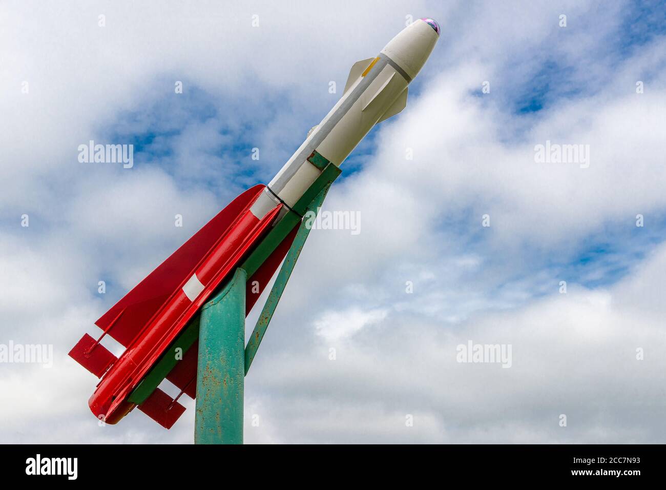 Eine kleine rote und weiße Rakete auf dem Display. Es ist auf einem Gier-Stand montiert und zeigt nach oben. Wolkiger Himmel vor dem Hintergrund mit einigen offenen Stellen. Stockfoto