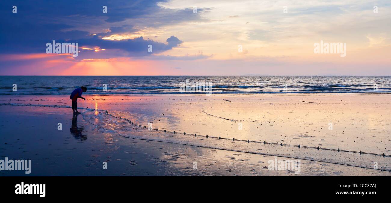Eine asiatische Fischer Frau fangen Salzwasserfische durch die Verwendung des Fischernetzes am Strand in der Dämmerung, dramatische Sonnenuntergang Himmel in den Hintergründen. Thailand. Stockfoto