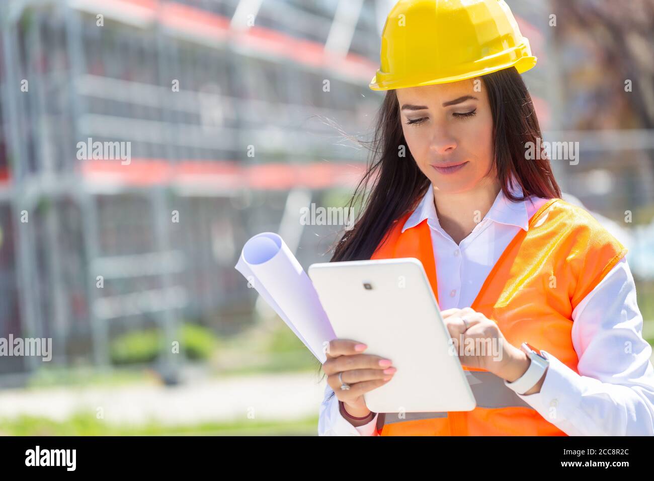Junge Frau in PSA hält eine Tablette und eine Blaupause mit einer Baustelle im Hintergrund. Stockfoto