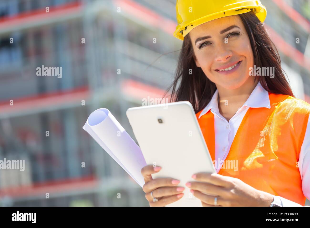Lächelnde Frau in PSA hält ein Tablet und eine Blaupause mit einer Baustelle im Hintergrund. Stockfoto