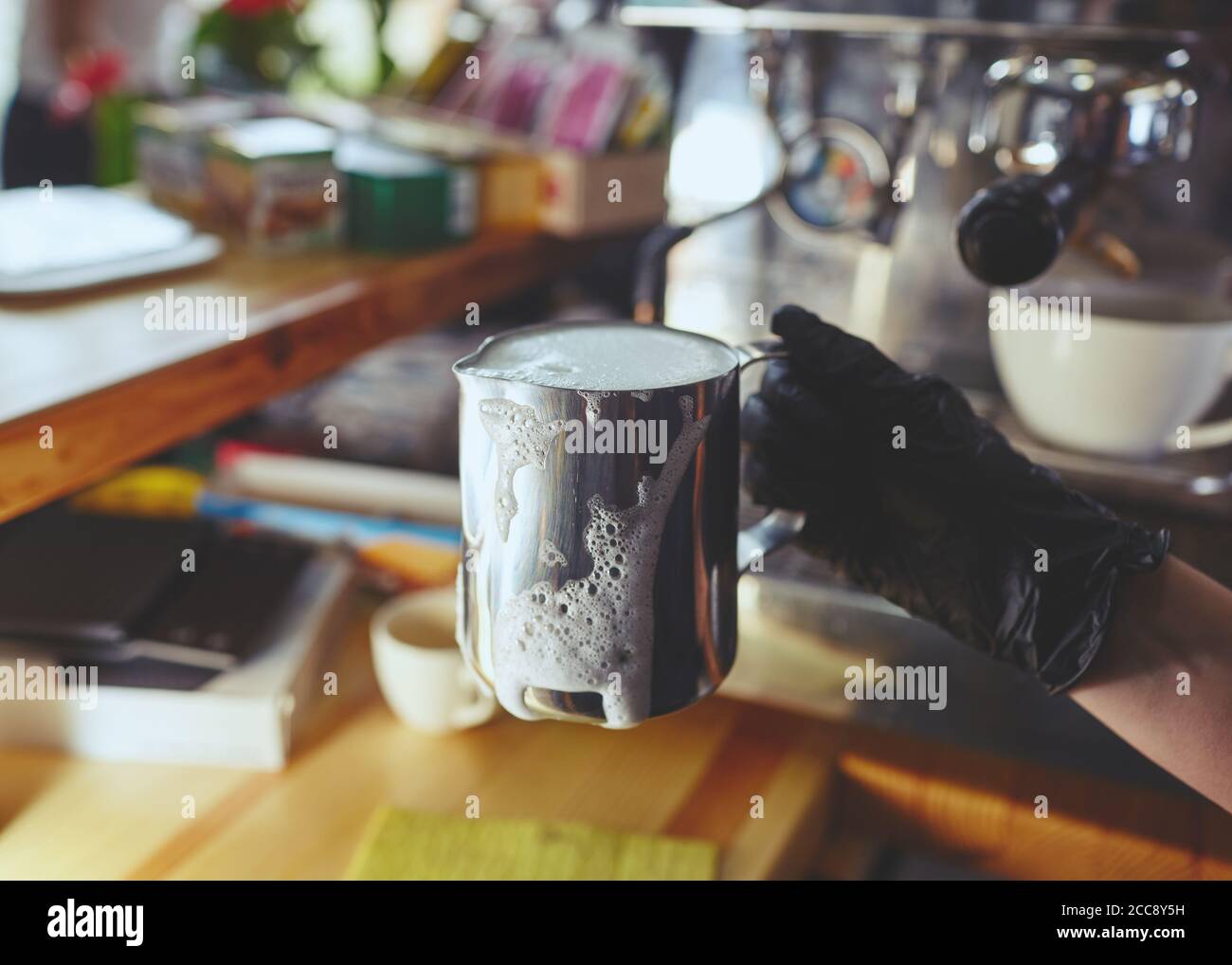Barista trägt medizinische Latexhandschuhe, bereitet Kaffeegetränk mit Espressomaschine im Shop Café zu. Frau hält den Krug mit Metallmilch in der Hand. Stockfoto