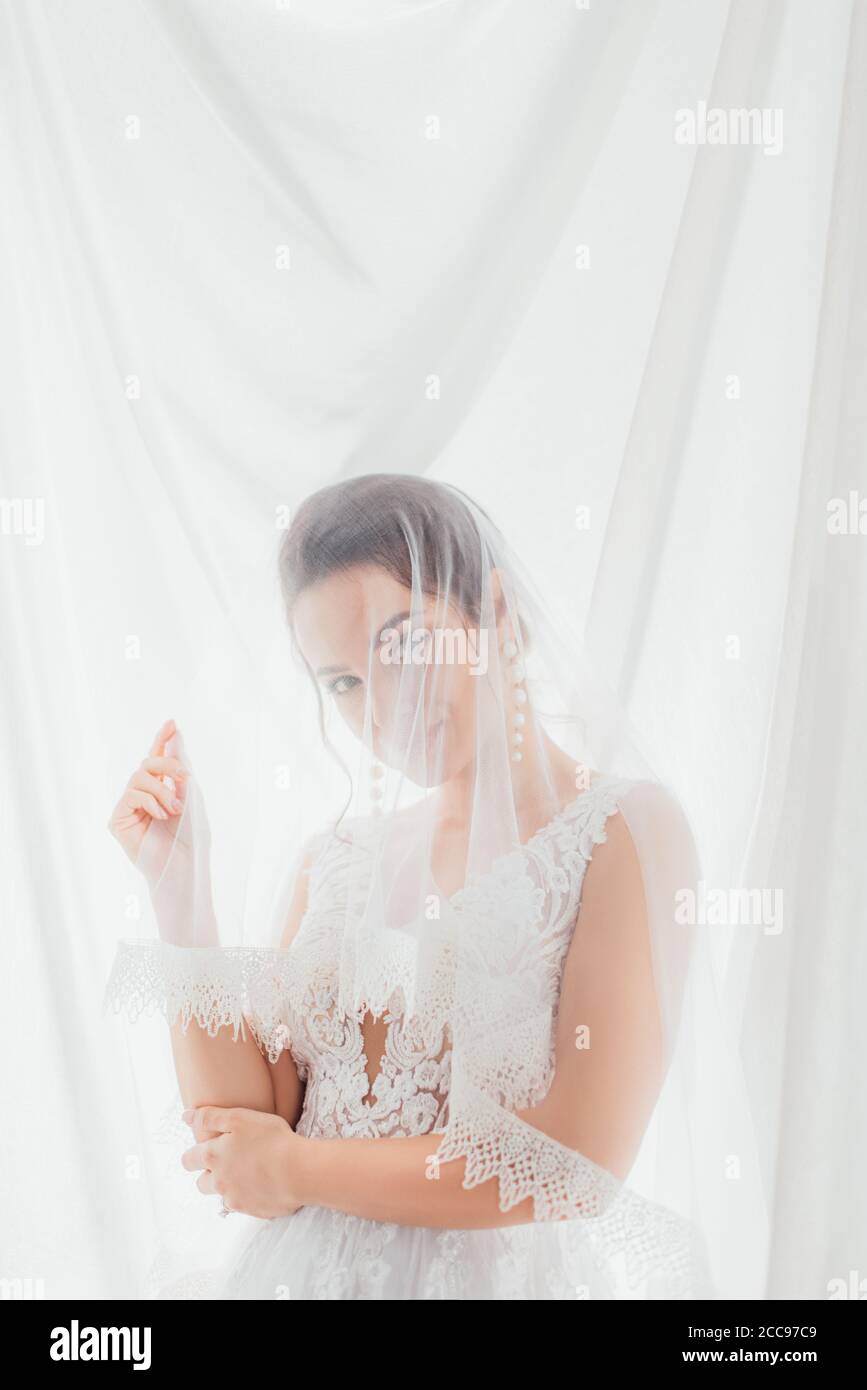Braut in Hochzeitskleid und Schleier Blick auf Kamera in der Nähe Weißes Tuch Stockfoto