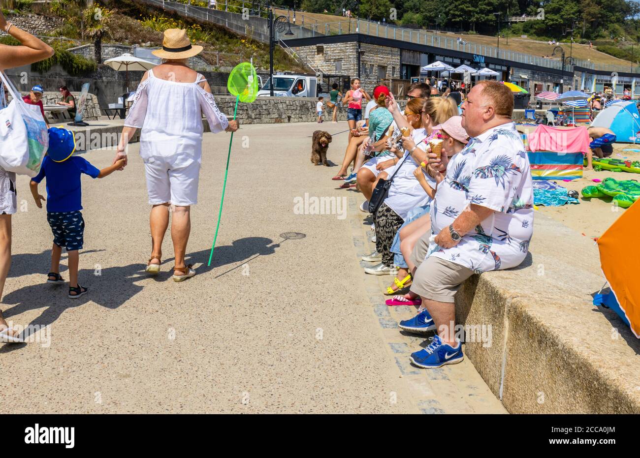 Übergewichtige Touristen sitzen an einer Strandmauer Essen Eiskühe, Lyme Regis, ein beliebter Badeort an der Jurassic Coast, Dorset, SW England Stockfoto