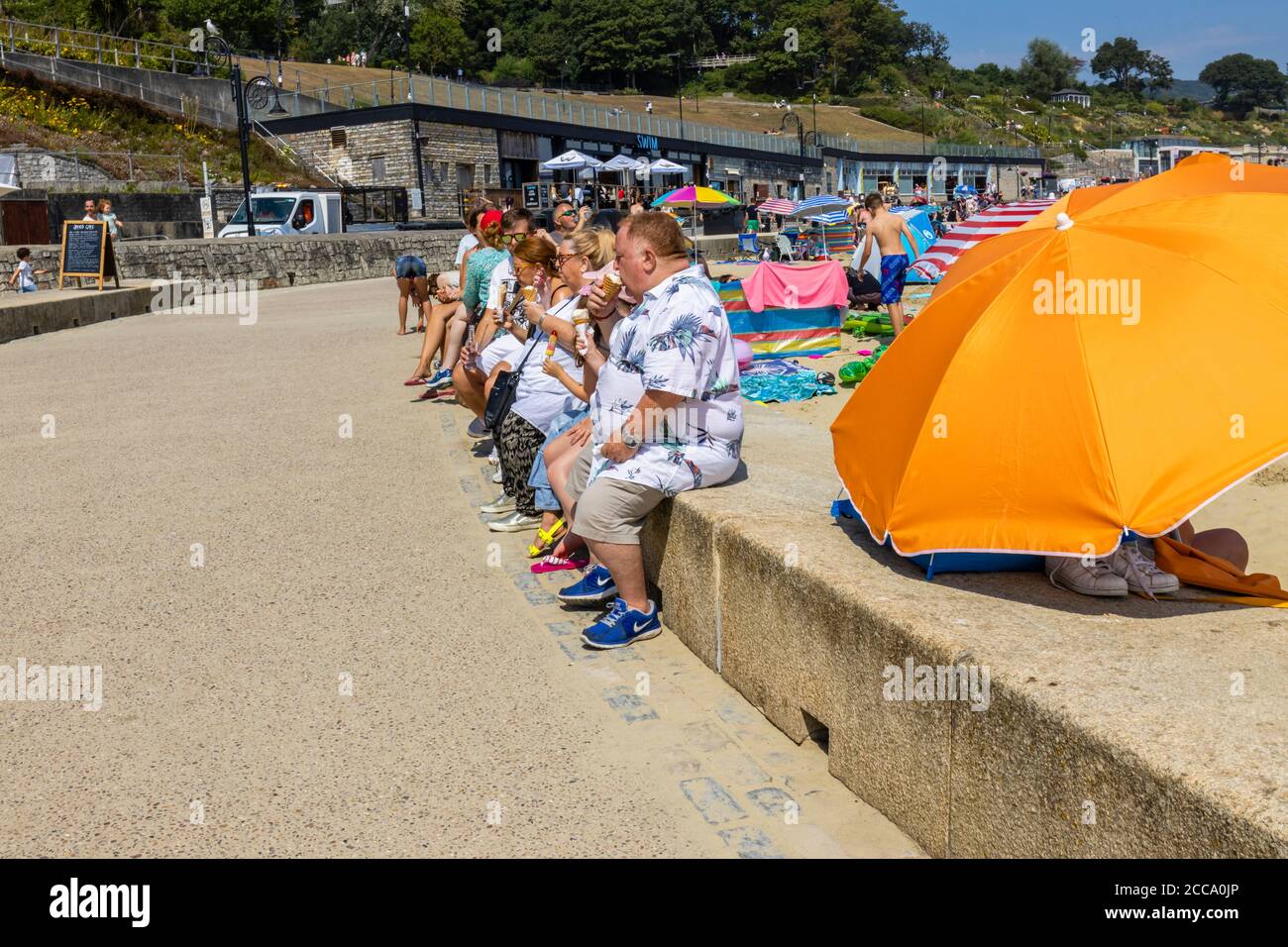 Übergewichtige Touristen sitzen an einer Strandmauer Essen Eiskühe, Lyme Regis, ein beliebter Badeort an der Jurassic Coast, Dorset, SW England Stockfoto