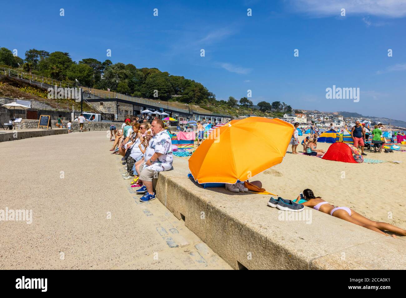 Übergewichtige Touristen sitzen an einer Strandmauer Essen Eiskühe, Lyme Regis, ein beliebter Badeort an der Jurassic Coast, Dorset, SW England Stockfoto