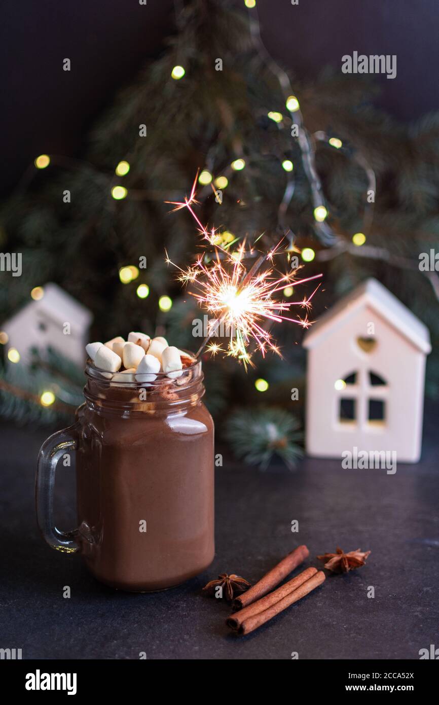 Heiße Schokolade im Glas-Einmachglas mit Marshmallows, Gewürzen und Sparkler auf schwarzem Tisch, umgeben von Tannenbaum, Weihnachtsbeleuchtung und Dekorationen. S Stockfoto