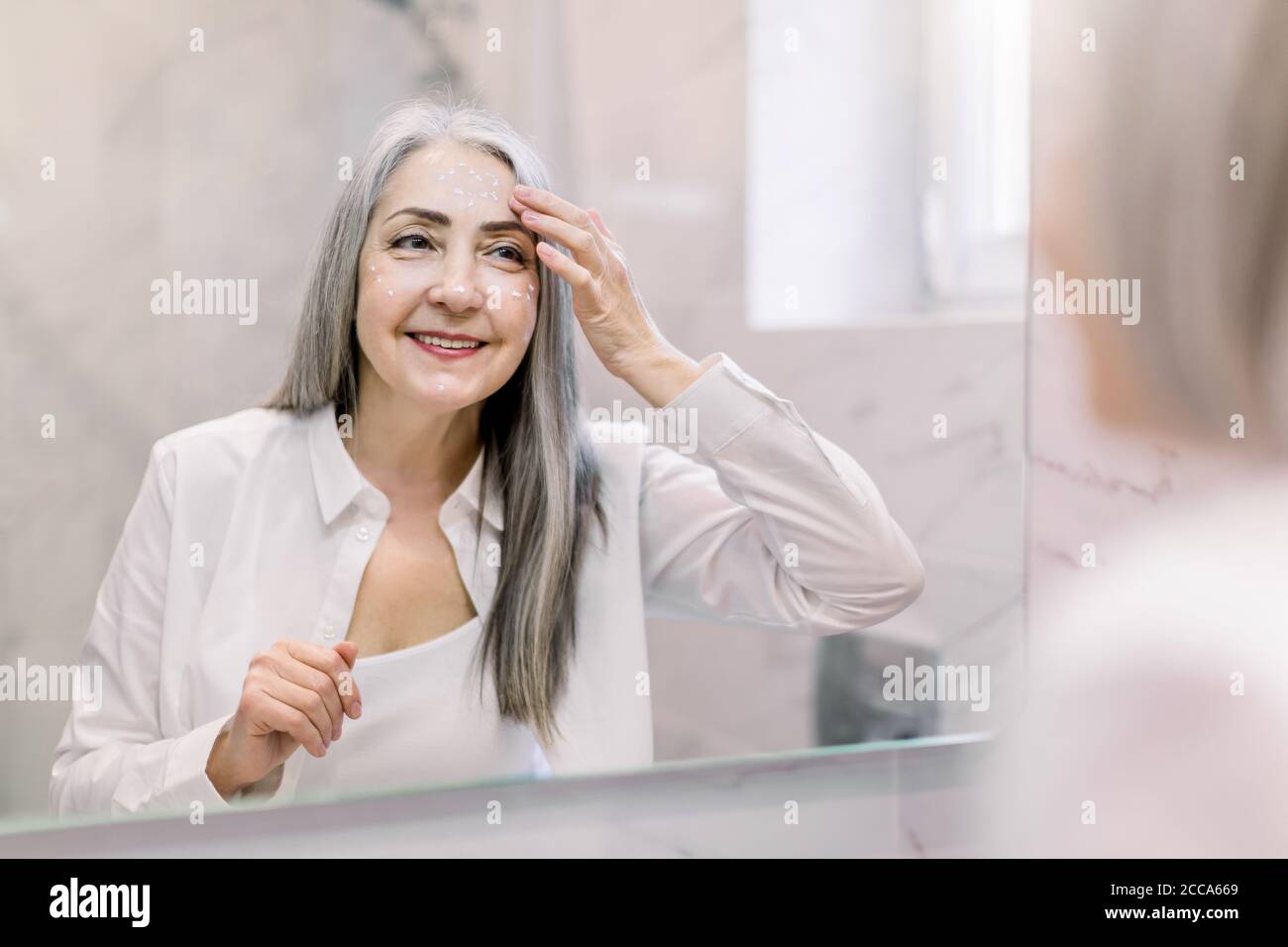 Hübsche ältere Frau mit langen grauen Haaren, trägt weißes Hemd, Blick auf ihr Gesicht im Badezimmerspiegel, und Anwendung Anti-Falten-Creme oder Kosmetik Stockfoto