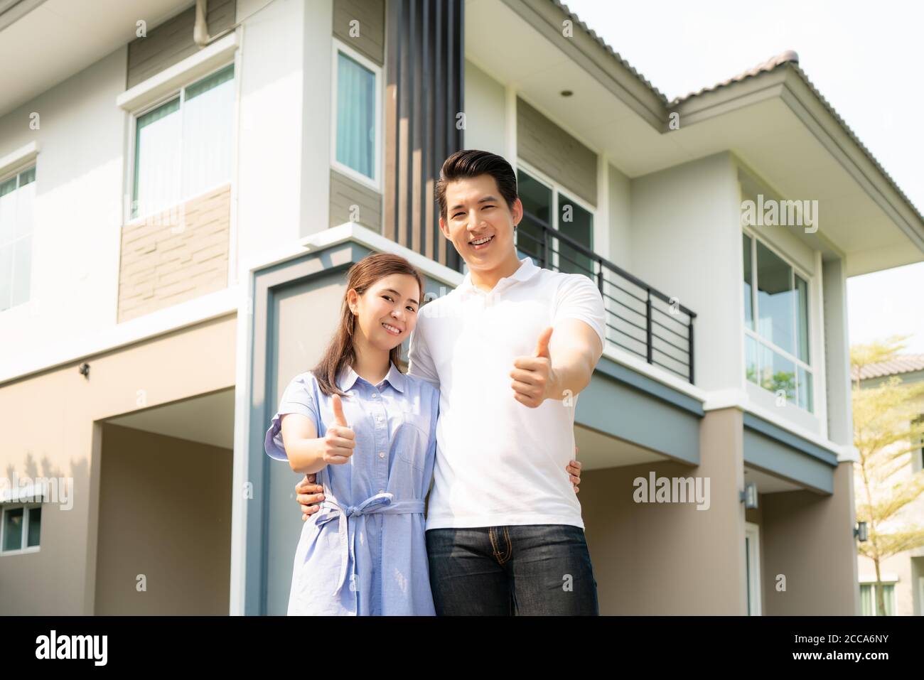 Portrait des asiatischen jungen Paares stehend, umarmt und zeigt Daumen nach oben zusammen glücklich vor ihrem neuen Haus, um neues Leben zu beginnen. Familie, ag Stockfoto