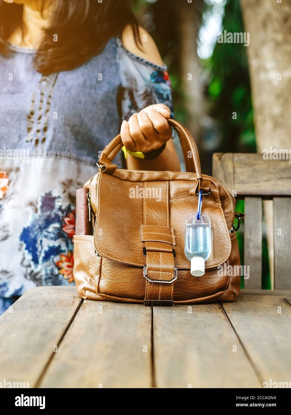 Mini tragbare Alkohol-Gel-Flasche zu töten Corona Virus (Covid-19) hängen auf einem braunen Leder Umhängetasche auf dem Tisch im Garten. Neuer normaler Lebensstil. Gesundheit Stockfoto