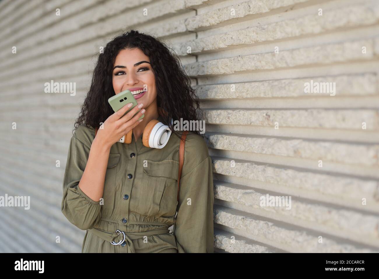 Junge Mixed Race Frau mit Kopfhörern in khaki Kleid mit Ihr Telefon in der Straße Stockfoto