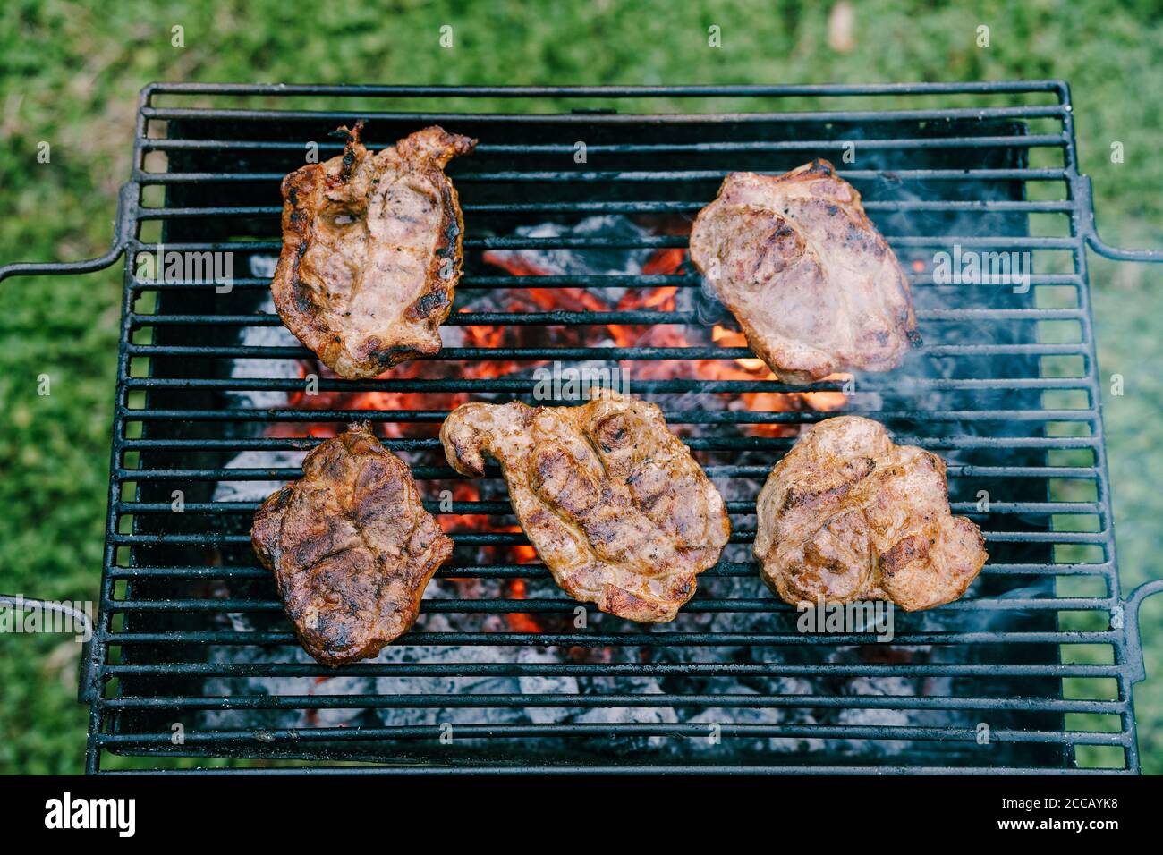 Fünf Fleischstücke auf einem schwarzen Metallgitter. Steaks werden auf heißen Kohlen gebraten, vor dem Hintergrund von Gras im Garten. Stockfoto