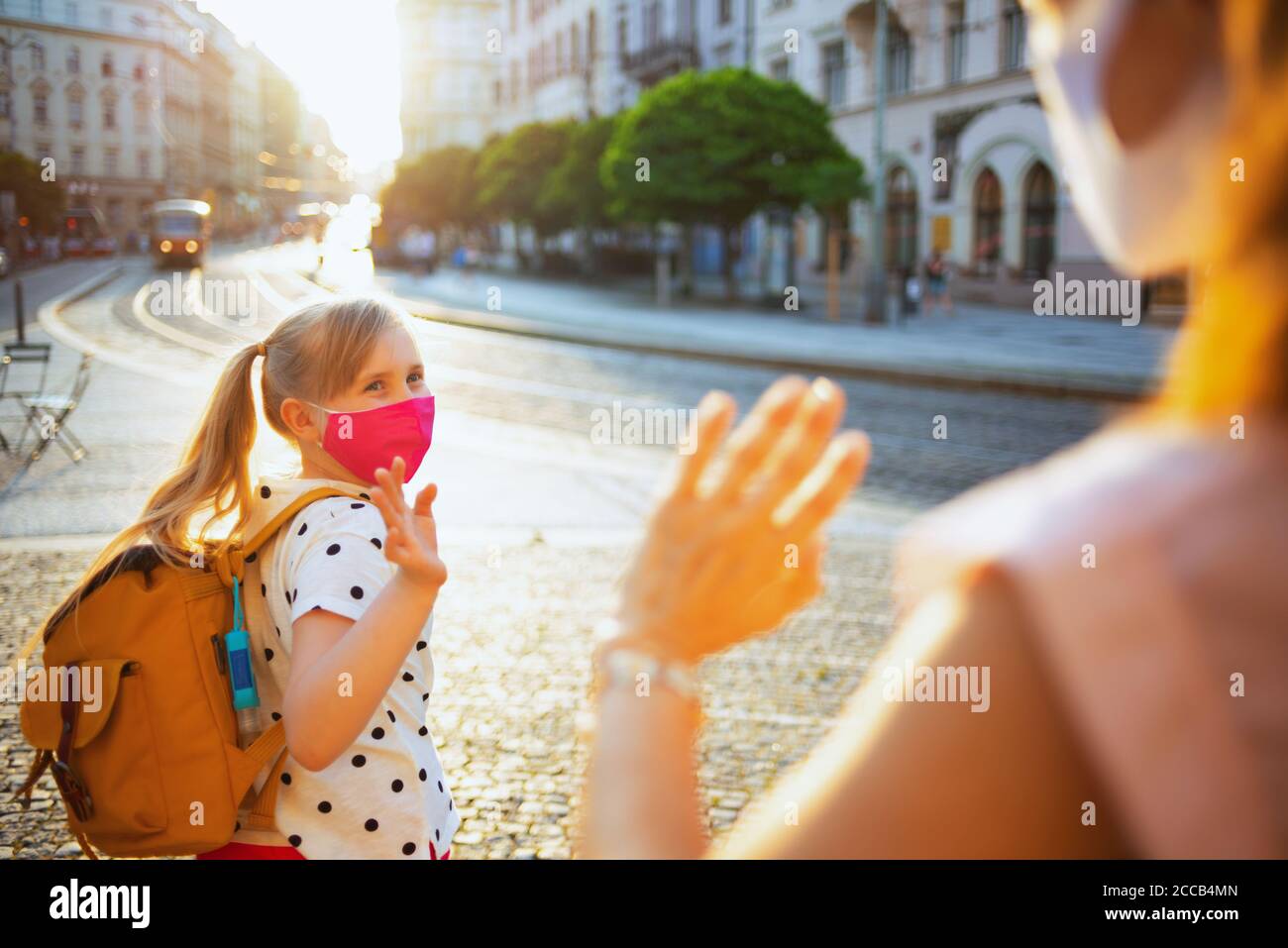 Leben während covid-19 Pandemie. Elegante Mutter und Kind mit Masken und gelben Rucksack sagen Auf Wiedersehen vor der Schule im Freien. Stockfoto