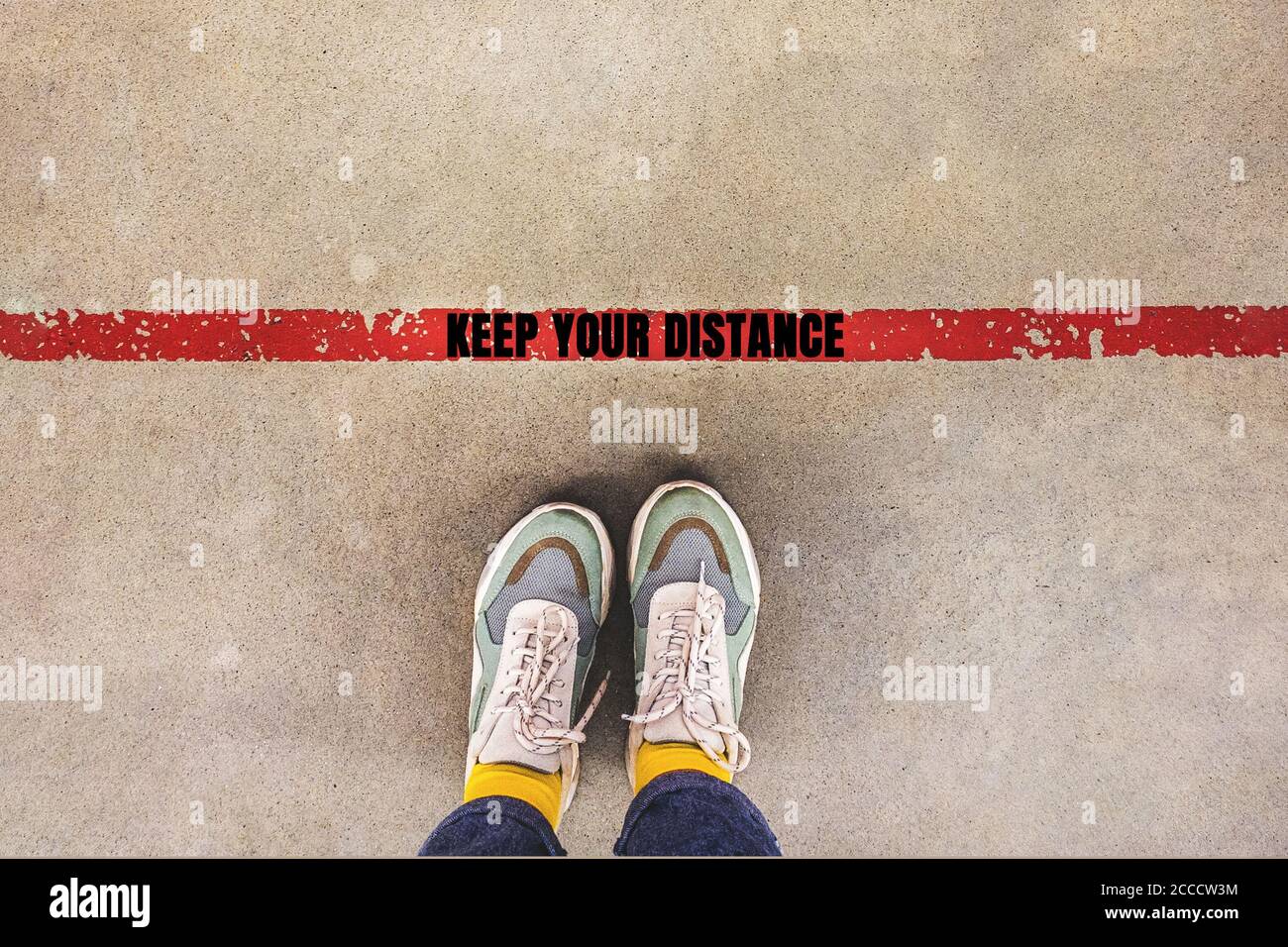 Person steht auf roter Linie mit Inschrift HALTEN SIE IHRE DISTANZ - soziale Distanzierung singen, als ein neuer normaler Lebenstrend. Sicherheitsabstand beibehalten Stockfoto
