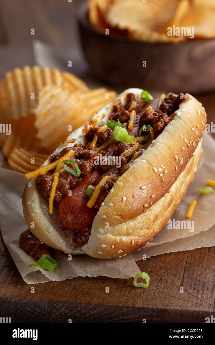 Chili Hot Dog mit Cheddar-Käse und grünen Zwiebeln auf Ein Sesambrötchen mit Kartoffelchips auf einem rustikalen Holzbrett Stockfoto