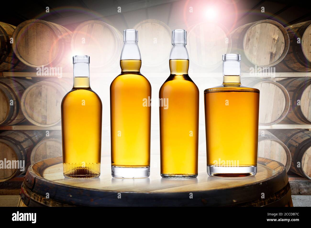 4 Flaschen glühender bernsteinfarbener Whisky, in verschiedenen Formen, auf einem alten Fass, in einem Fasslager, hinterleuchtet mit buntem Linsengeflare. Stockfoto