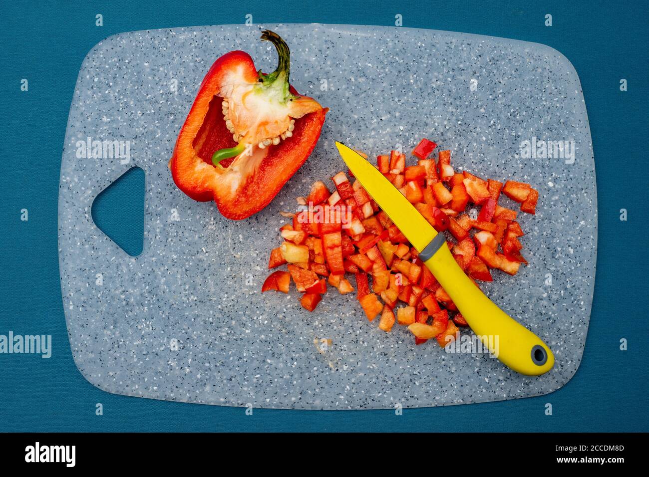 Draufsicht auf einen halbierten und gehackten roten Pfeffer mit einem gelben Messer auf einem blauen Brett und blauem Hintergrund. Stockfoto