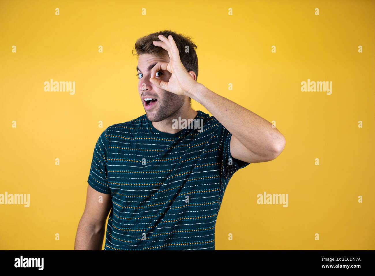 Hübscher Mann, der über isoliertem gelben Hintergrund steht, tut okay Geste schockiert mit überraschendem Gesicht, Auge schaut durch die Finger Stockfoto