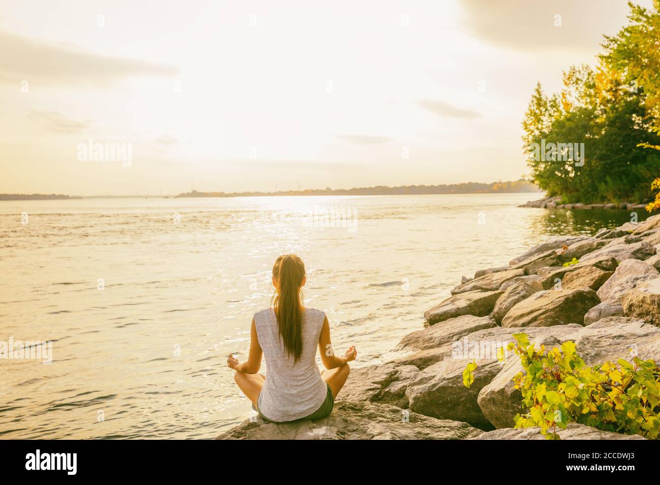 Yoga-Kurs im Freien im Naturpark am Ufer des Sees. Frau in Lotushaltung sitzend meditiert am Wasser in Morgensonne aufgehen. Meditation Stockfoto